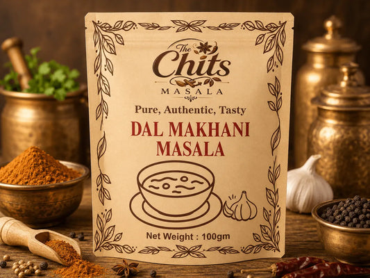 A package of Dal Makhani Masala by The Chits Masala stands upright on a rustic wooden surface, surrounded by spices, brass containers, garlic, and dried red chilies in a warm, appetizing setting.