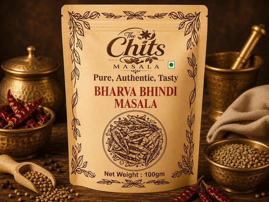 A 100g stand-up pouch of 'The Chits Masala' Bharya Bindi Masala centered on a rustic wooden table surrounded by ingredients like whole red chilies, coriander seeds, and brass bowls.
