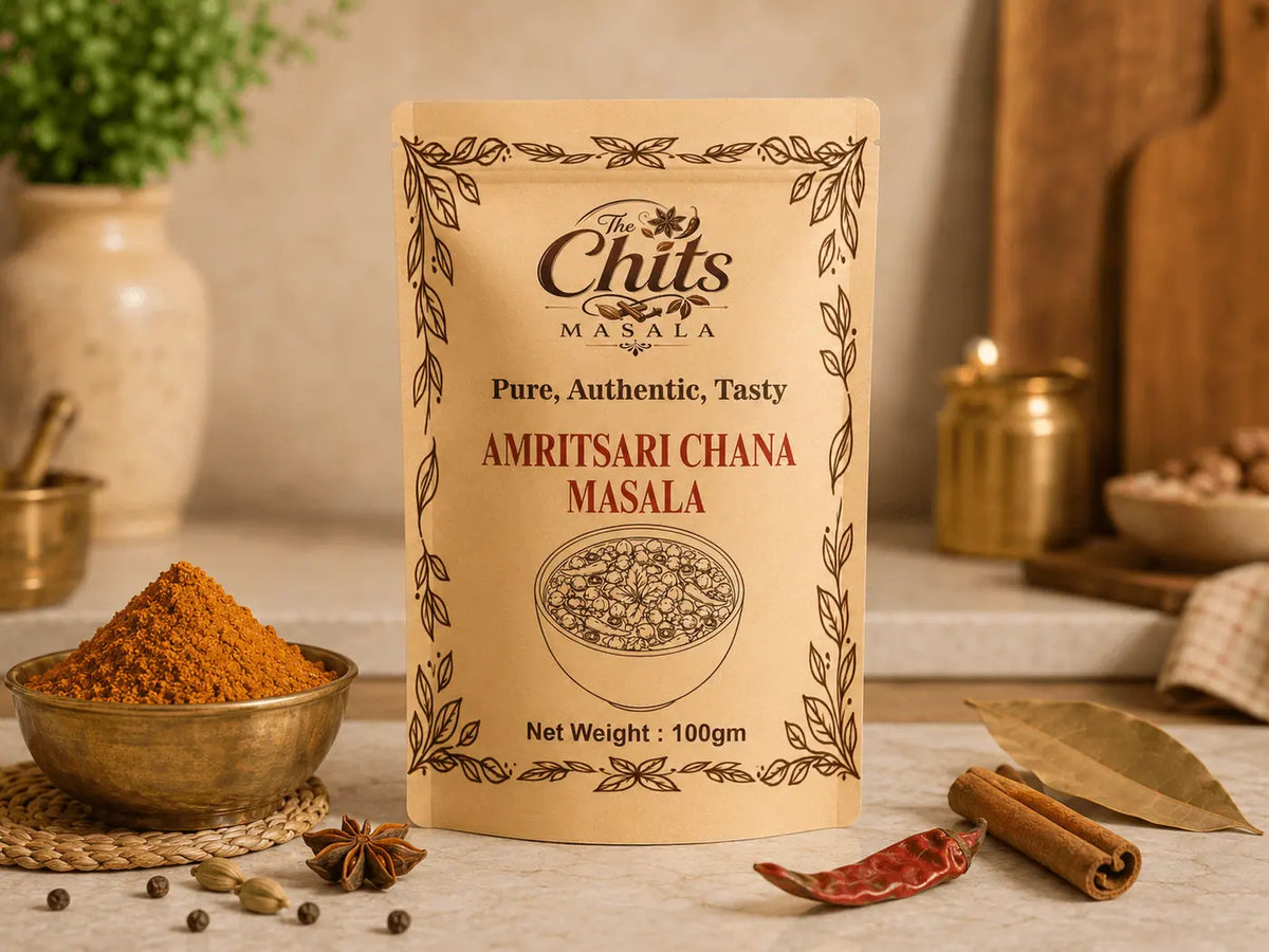 A 100g stand-up pouch of Amritsari Chana Masala spice mix by The Chits Masala, styled on a marble countertop with a bowl of orange-red chili powder, cinnamon sticks, and bay leaves in a warm kitchen setting.