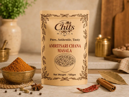 A 100g stand-up pouch of Amritsari Chana Masala spice mix by The Chits Masala, styled on a marble countertop with a bowl of orange-red chili powder, cinnamon sticks, and bay leaves in a warm kitchen setting.