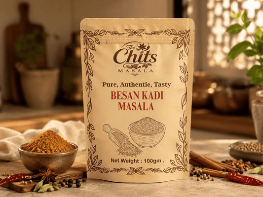 A 100g stand-up pouch of The Chits Masala Besan Kadi Masala spice mix, displayed on a rustic wooden table with bowls of the ground spice and whole spices like cinnamon and red chilies in a warm, softly blurred kitchen setting.
