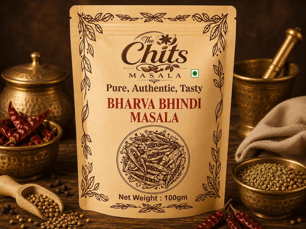 A 100g stand-up pouch of 'The Chits Masala' Bharya Bindi Masala centered on a rustic wooden table surrounded by ingredients like whole red chilies, coriander seeds, and brass bowls.