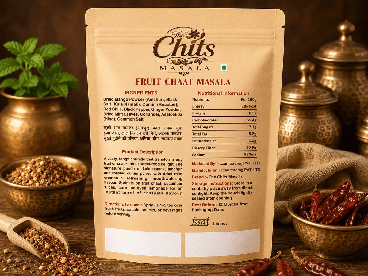 A stand-up pouch of The Chuts Masala Fruit Chaat Masala shown from the back, with product details, ingredients, and nutritional information printed in English and Hindi. The pouch is styled on a dark wooden surface with dried red chilies, a wooden spoon of spices, and brass containers in the background.