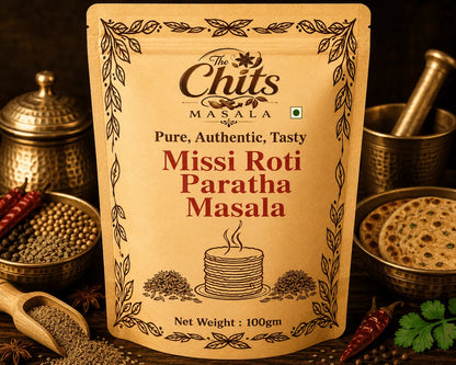 A stand-up pouch of The Chits Masala Missi Roti Paratha Masala seasoning, displayed in a styled flat-lay arrangement with brass bowls of whole spices, red chilies, a wooden spoon, and a cooked paratha on a dark background.